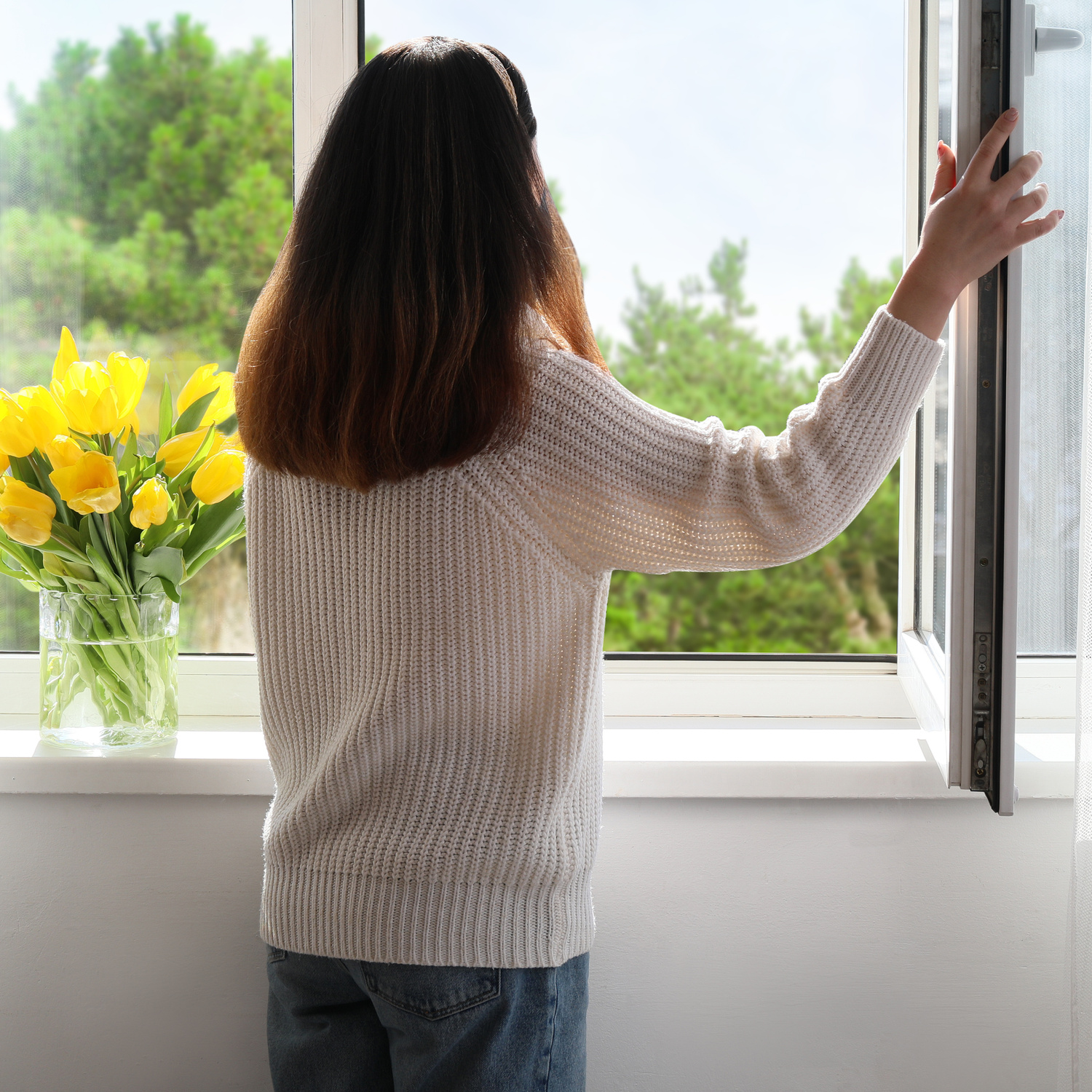 A young woman with her back to the camera, opening a window in her home and staring at the trees outside.