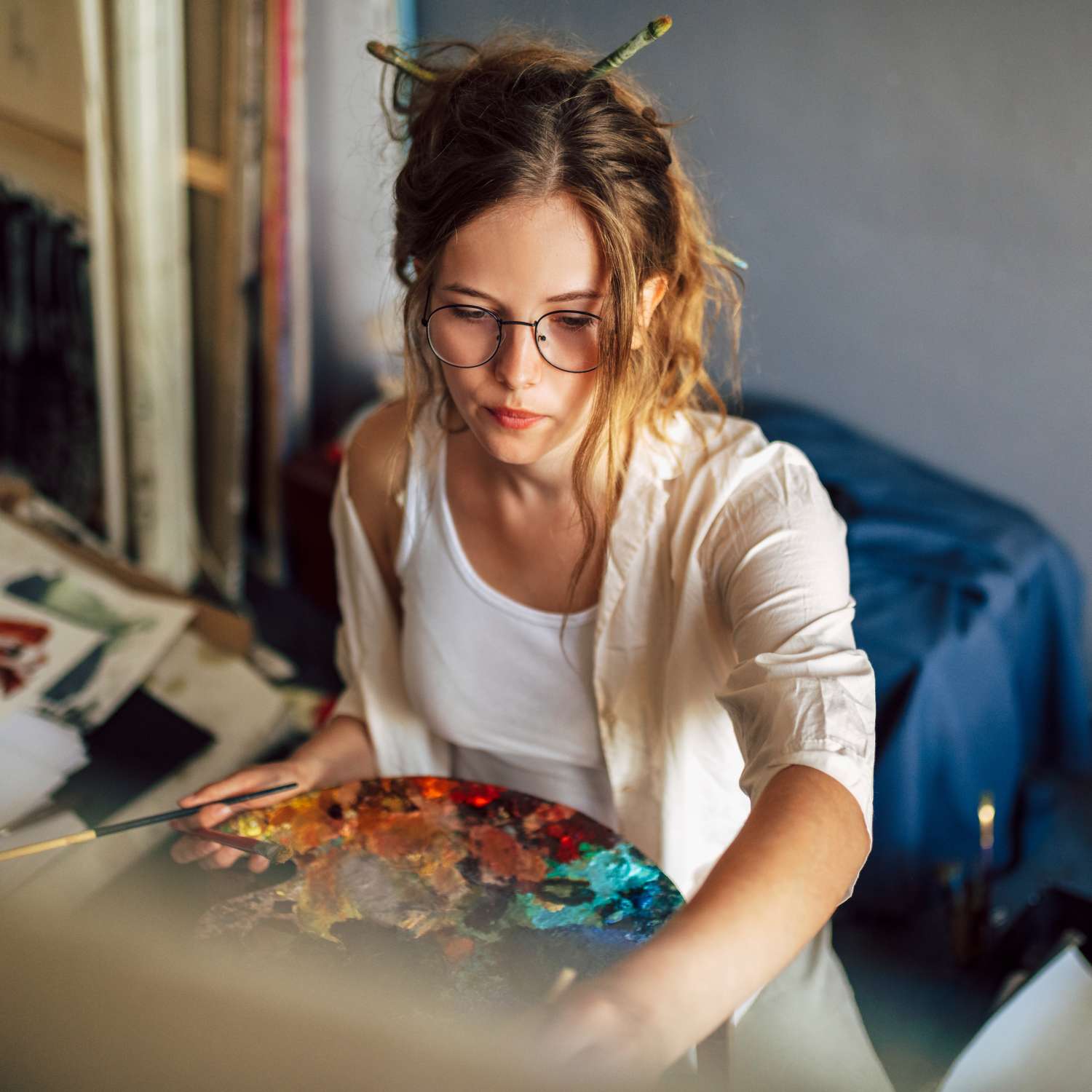 A woman at her easel working on a painting. She's wearing glasses with her hair pulled back and wearing a smock.