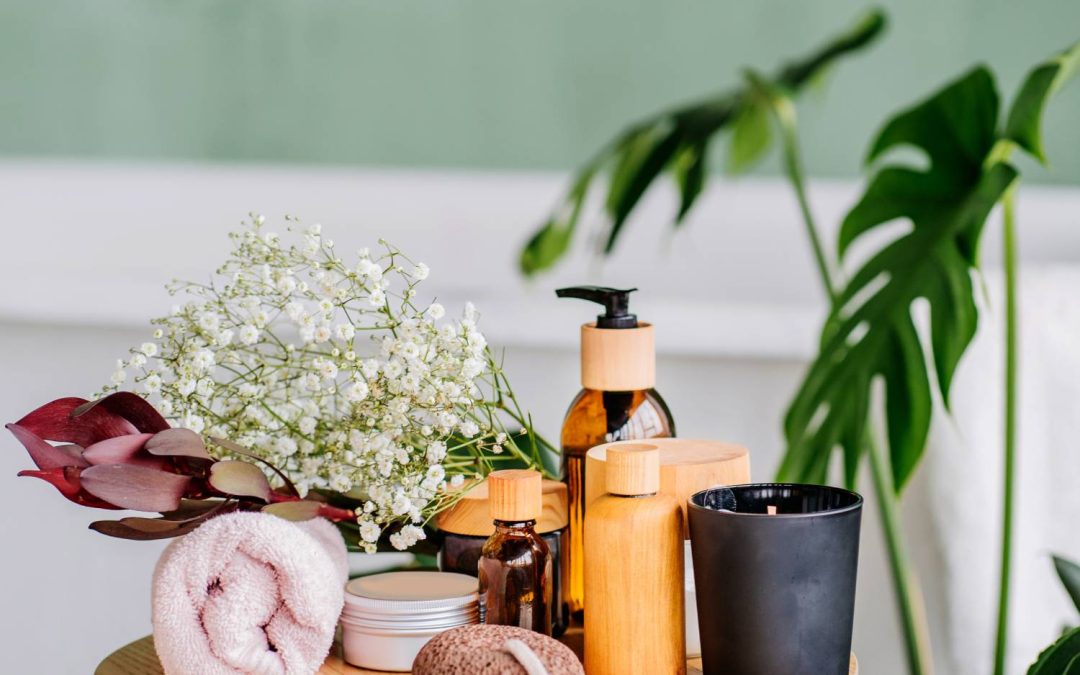 A side table with spa supplies, including a towel, bottled products, and pumice, with a bathtub and monstera in the background.