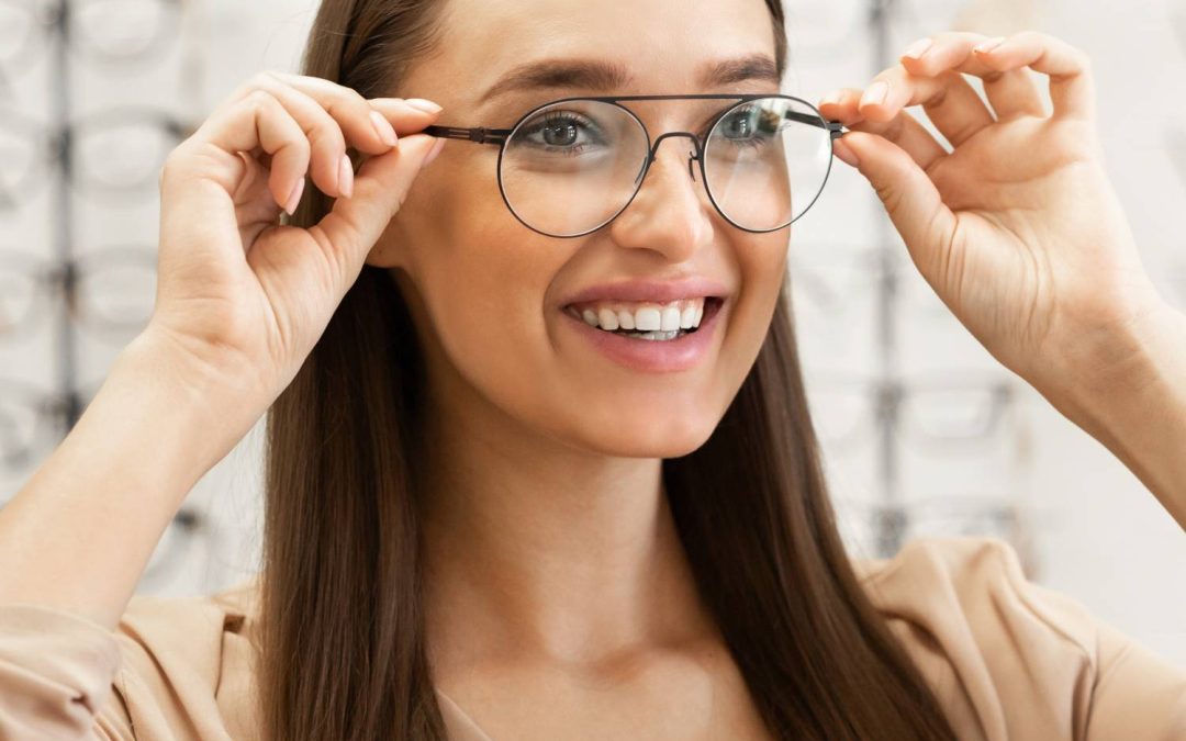 A young woman wearing a tan shirt is trying on a pair of frames. There are frames on display in the background.