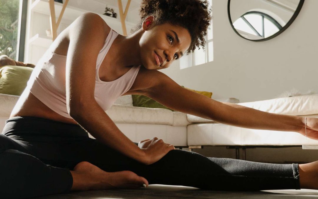 A young woman sits on a black yoga mat stretching. She's wearing black yoga pants and a light-pink sports bra.