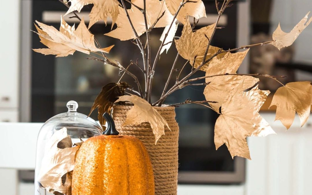 An orange glitter pumpkin and a vase filled with branches with gold leaves sit on a tray in the middle of a white table.