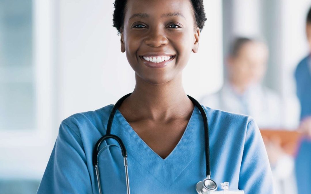 A Black, smiling nursing professional standing in the hallway with her arms folded. She is happy and wearing a stethoscope.