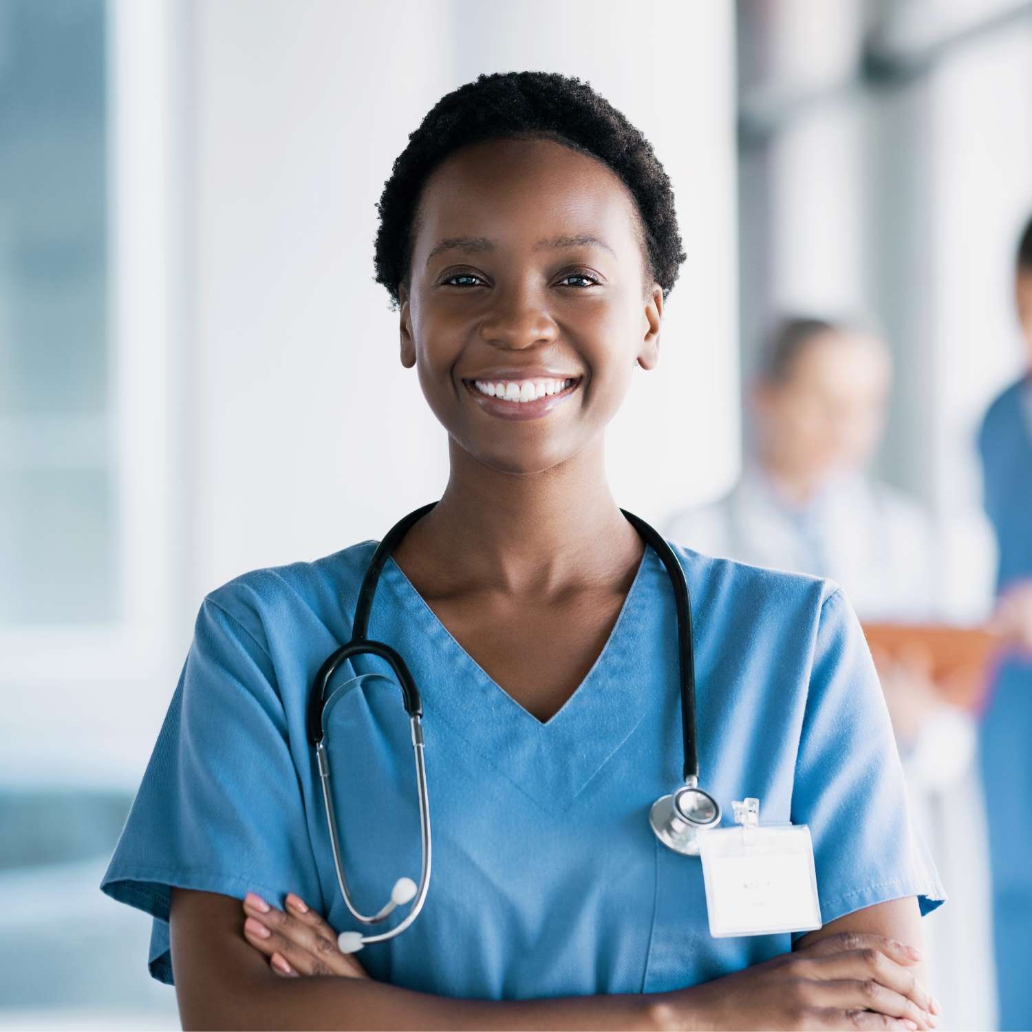 A Black, smiling nursing professional standing in the hallway with her arms folded. She is happy and wearing a stethoscope.